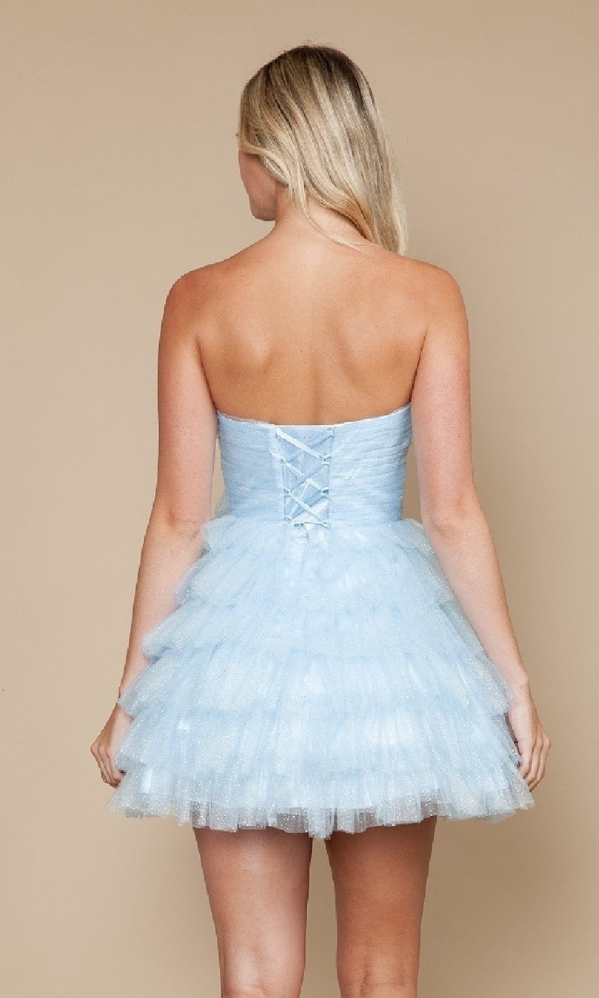 A woman with long blonde hair models the Strapless Short Tulle Hoco Dress: Poly USA 9912 in light blue, featuring a lace-up back and tiered A-line skirt, against a plain beige background.